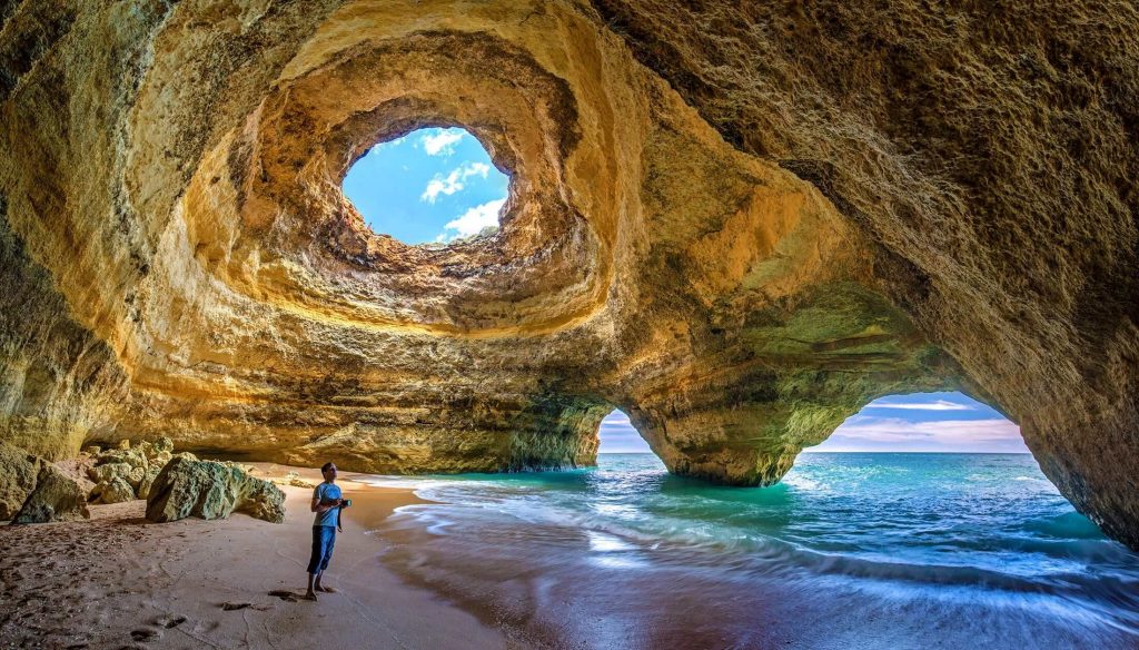 A view from the beach inside Benagil cave with a person standing on the beach observing the limestone walls, with a lookout towards the ocean and the 'Eye of Benagil' above.